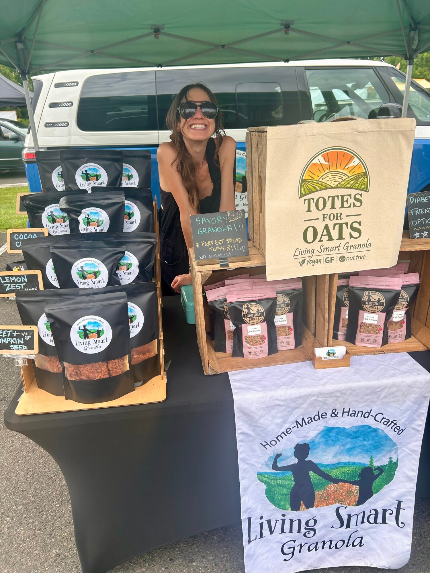 Woman smiling at a market stall with oat products and granola bags, under a green canopy, in front of a vehicle.