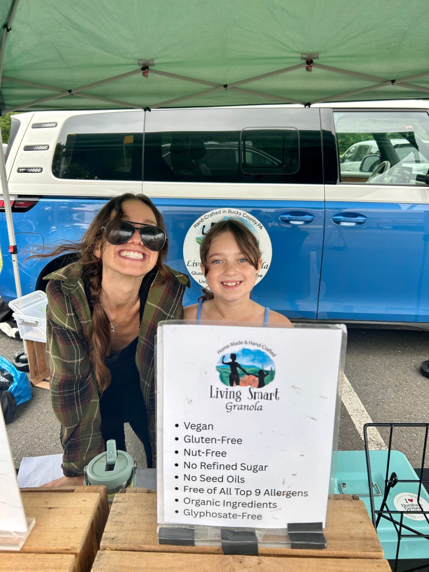 Two smiling people, a woman and a girl, sit at a table with a sign in front of them, outdoors near a blue and white vehicle.