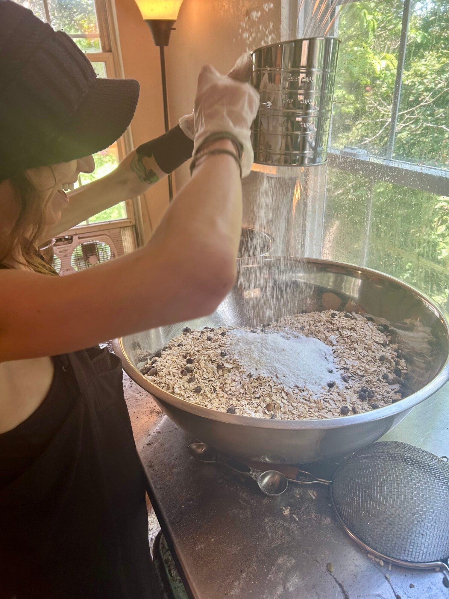 Person pouring dry ingredients into a large mixing bowl on a kitchen counter, with a window showing trees outside.