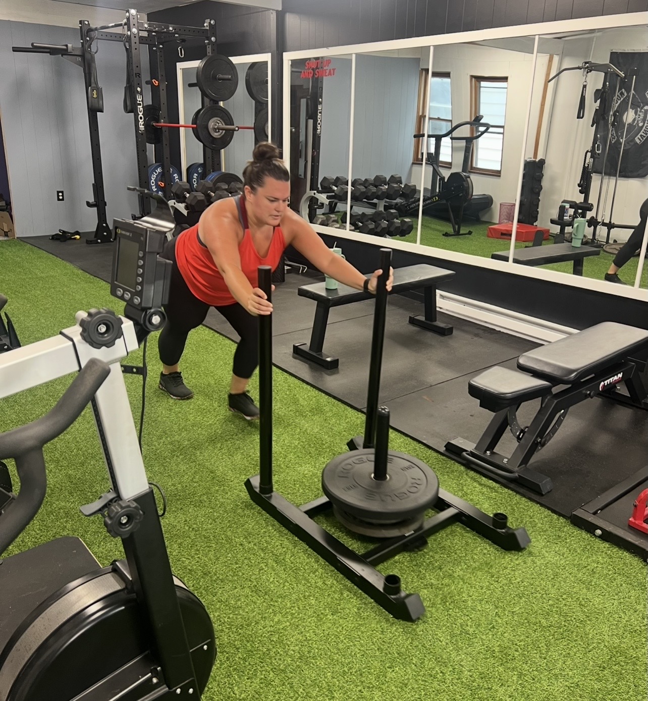 Woman in red top pushes a weighted sled in a gym with mirrors, weight racks, and exercise equipment, on green turf flooring.