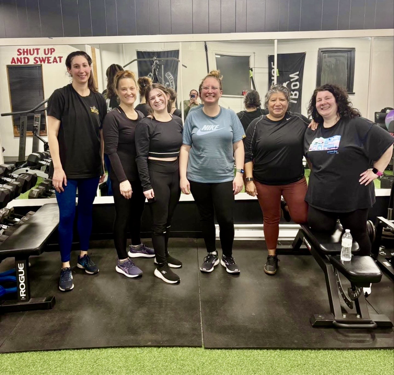 Six women standing in a gym, smiling, with workout equipment and mirrors behind them.