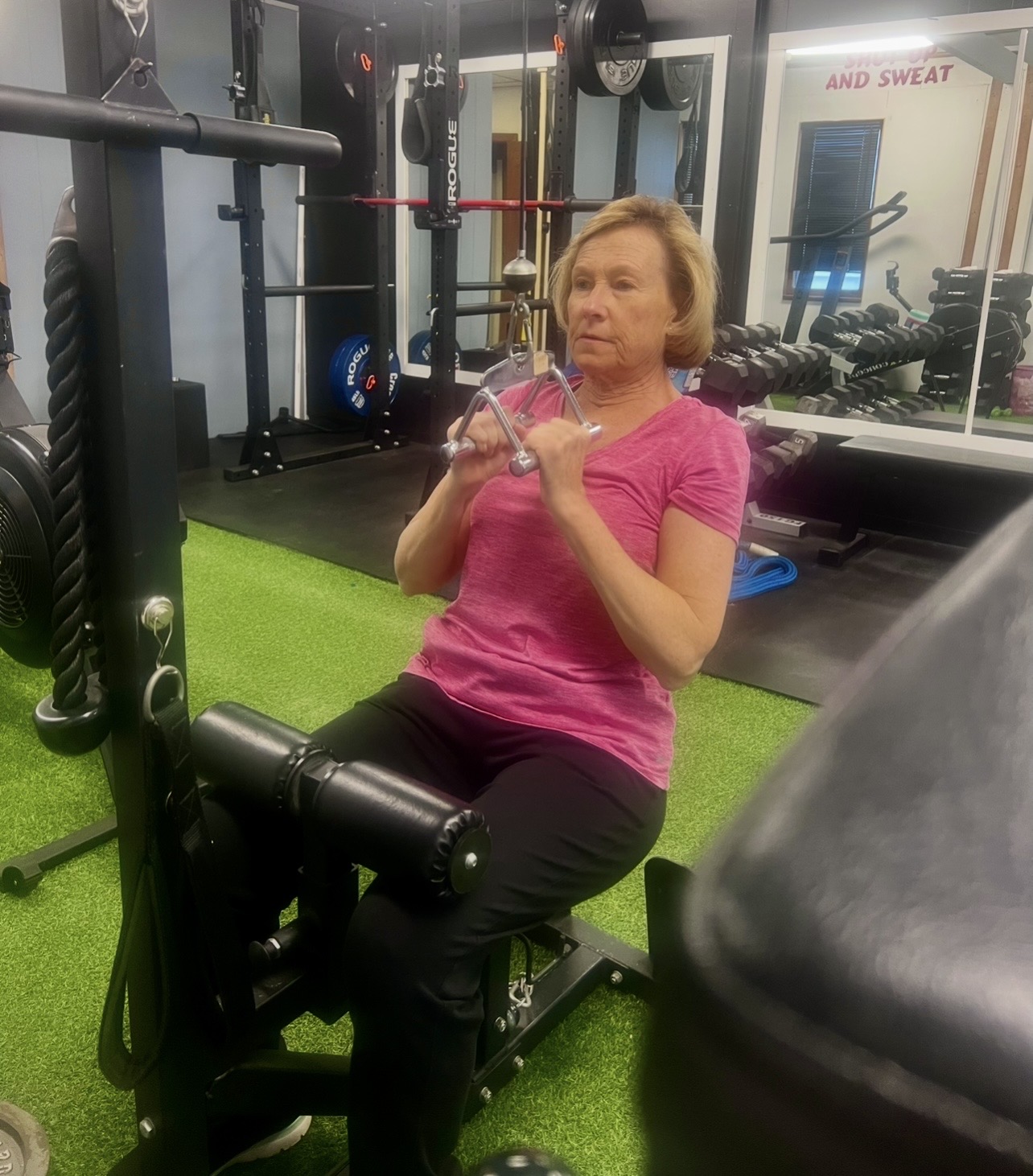 Woman in pink shirt sitting on a workout bench in a gym, holding a phone, with gym equipment in background.