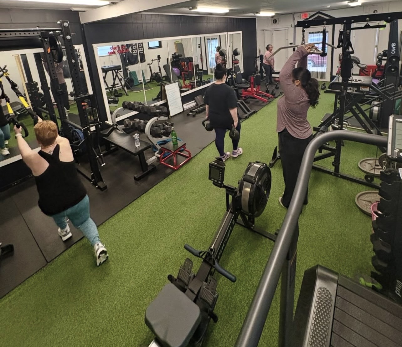People exercising in a gym with equipment, mirrors, and green turf flooring.
