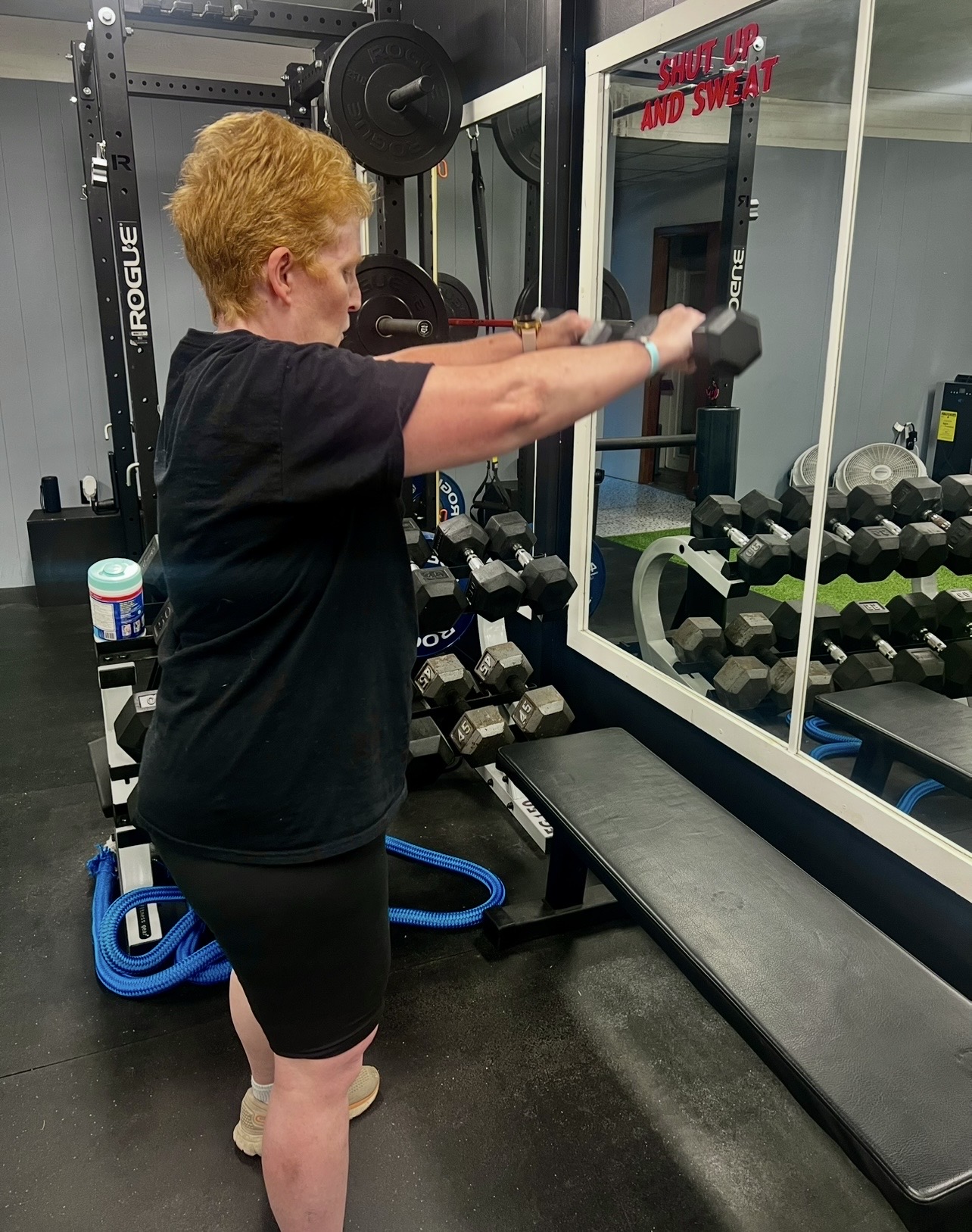 Person with short hair and black clothing stretching arms in a gym, with weights and mirrors in the background.