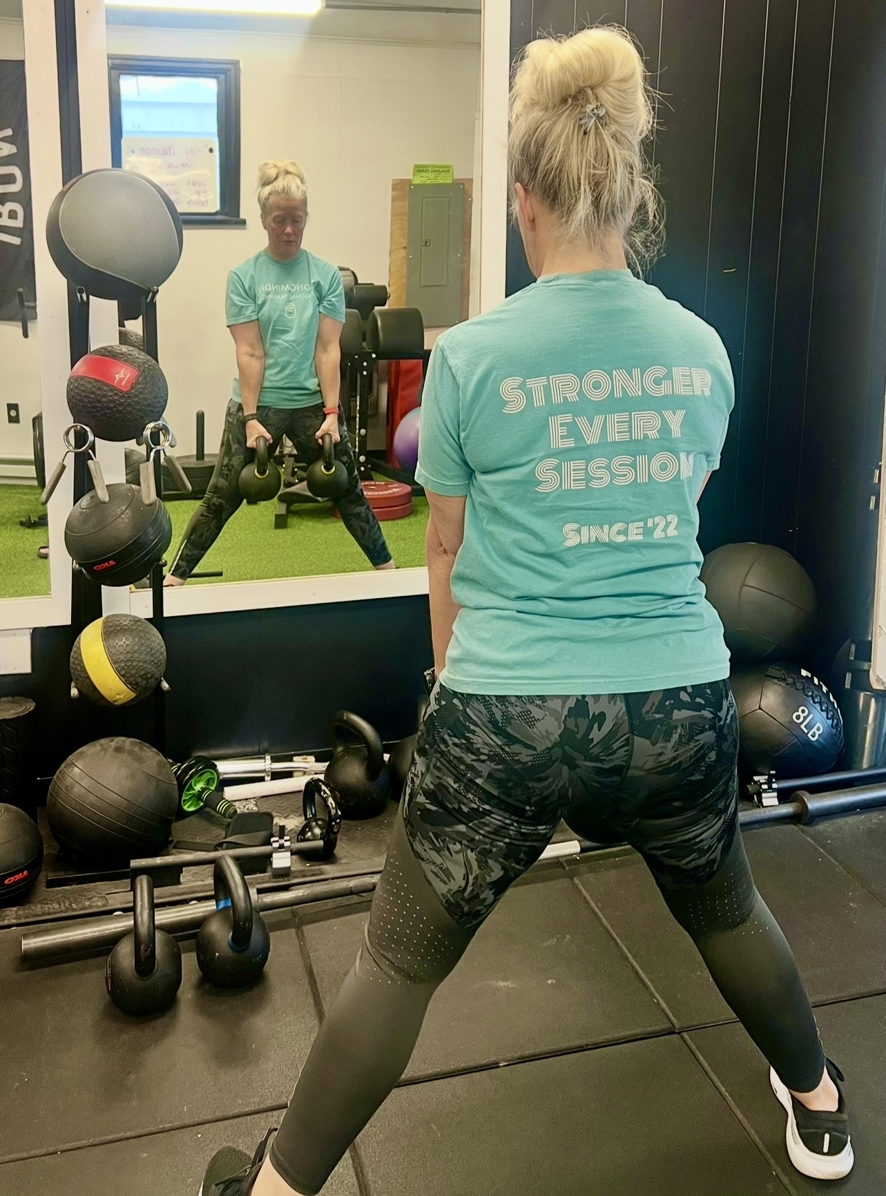 Woman with blonde hair in a bun wearing a turquoise shirt and patterned leggings exercises in front of a mirror, with a young girl visible in the reflection. Gym equipment around them.