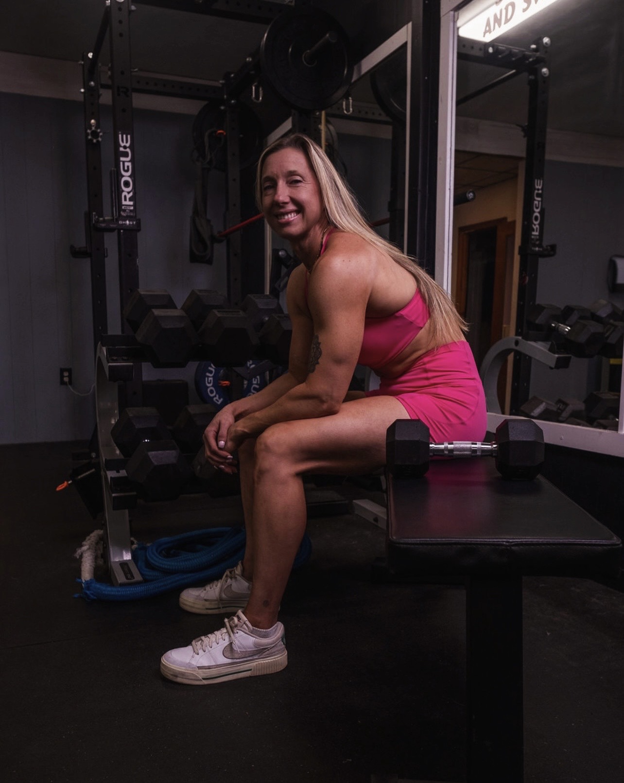 Woman in pink workout outfit sitting on a bench in a gym, smiling, with gym equipment around her.