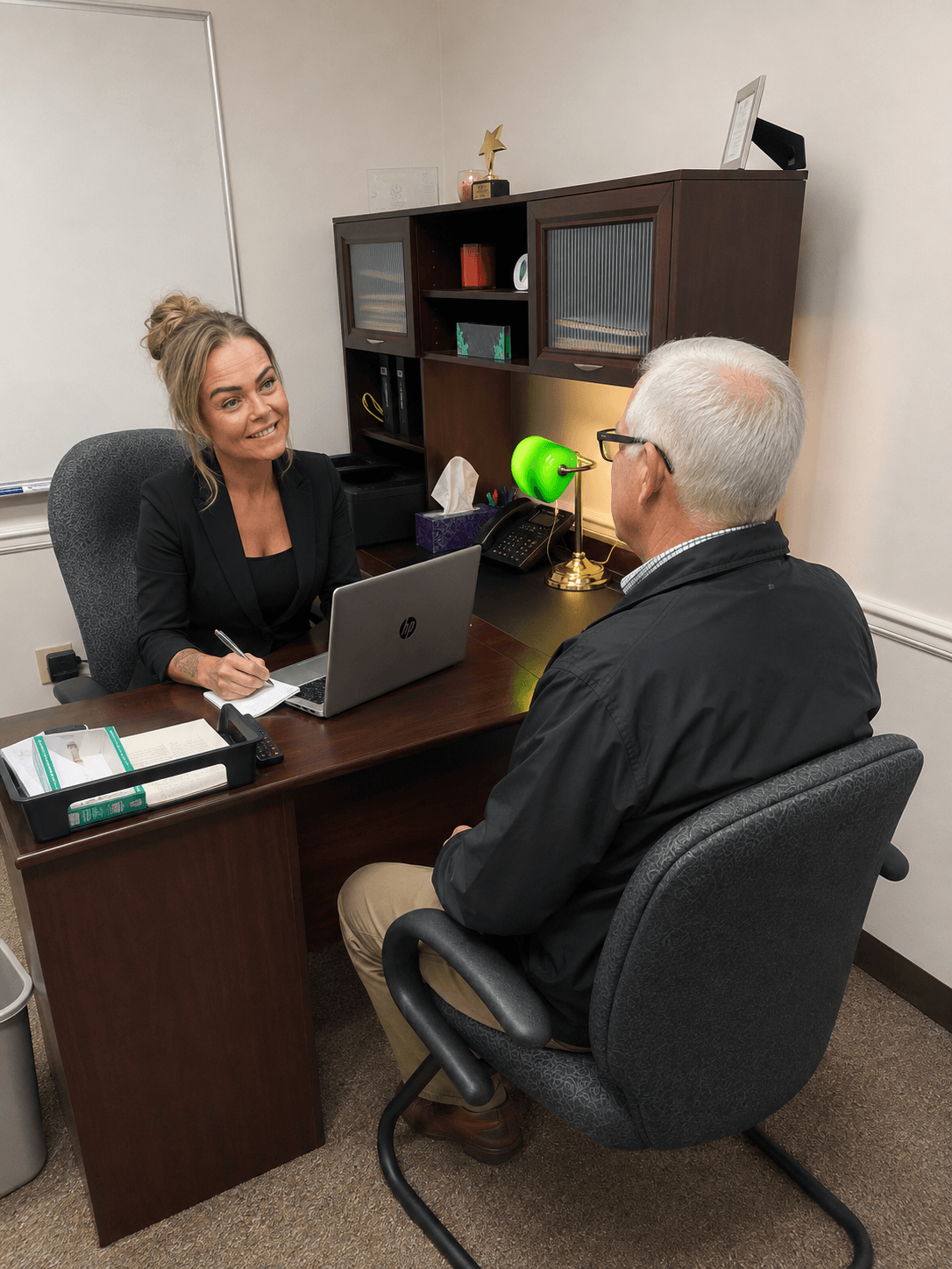 Woman and man sitting at a desk in an office, engaging in conversation, with a laptop and office supplies present.
