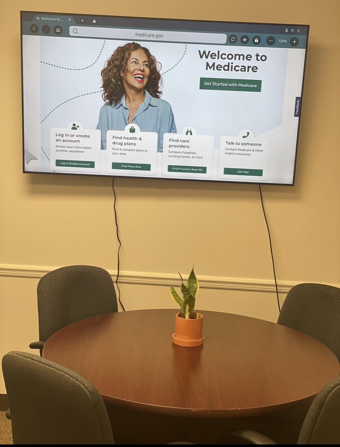 Conference room with a round table, four chairs, a potted plant, and a wall-mounted screen displaying Medicare website.