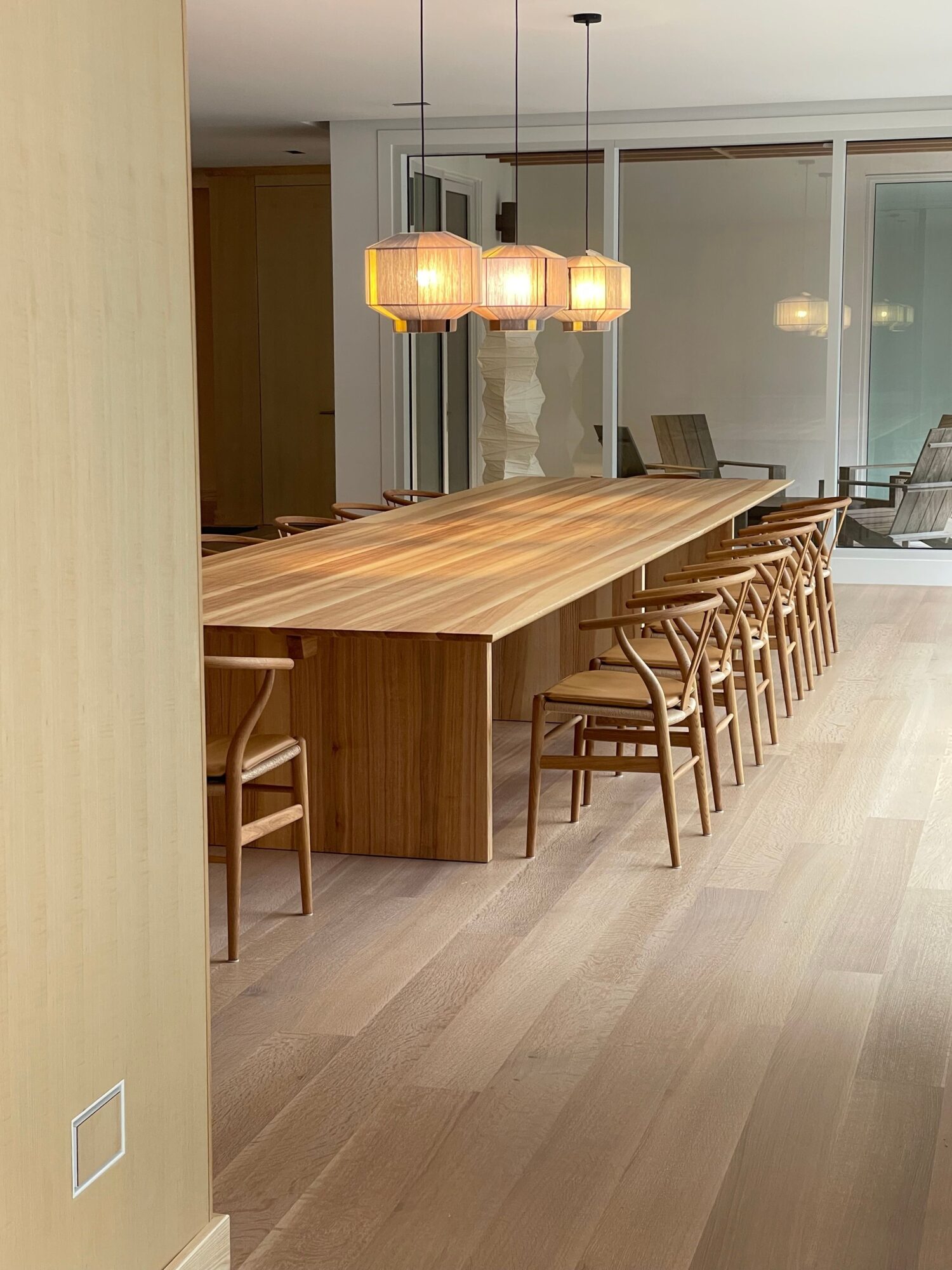 Long wooden dining table with chairs in a modern room, pendant lights hanging above, large windows in the background.