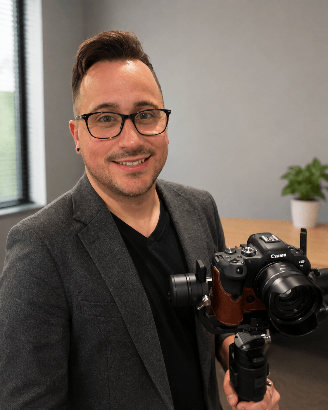 Man with glasses and a blazer holding a camera, smiling indoors near a window and a potted plant.