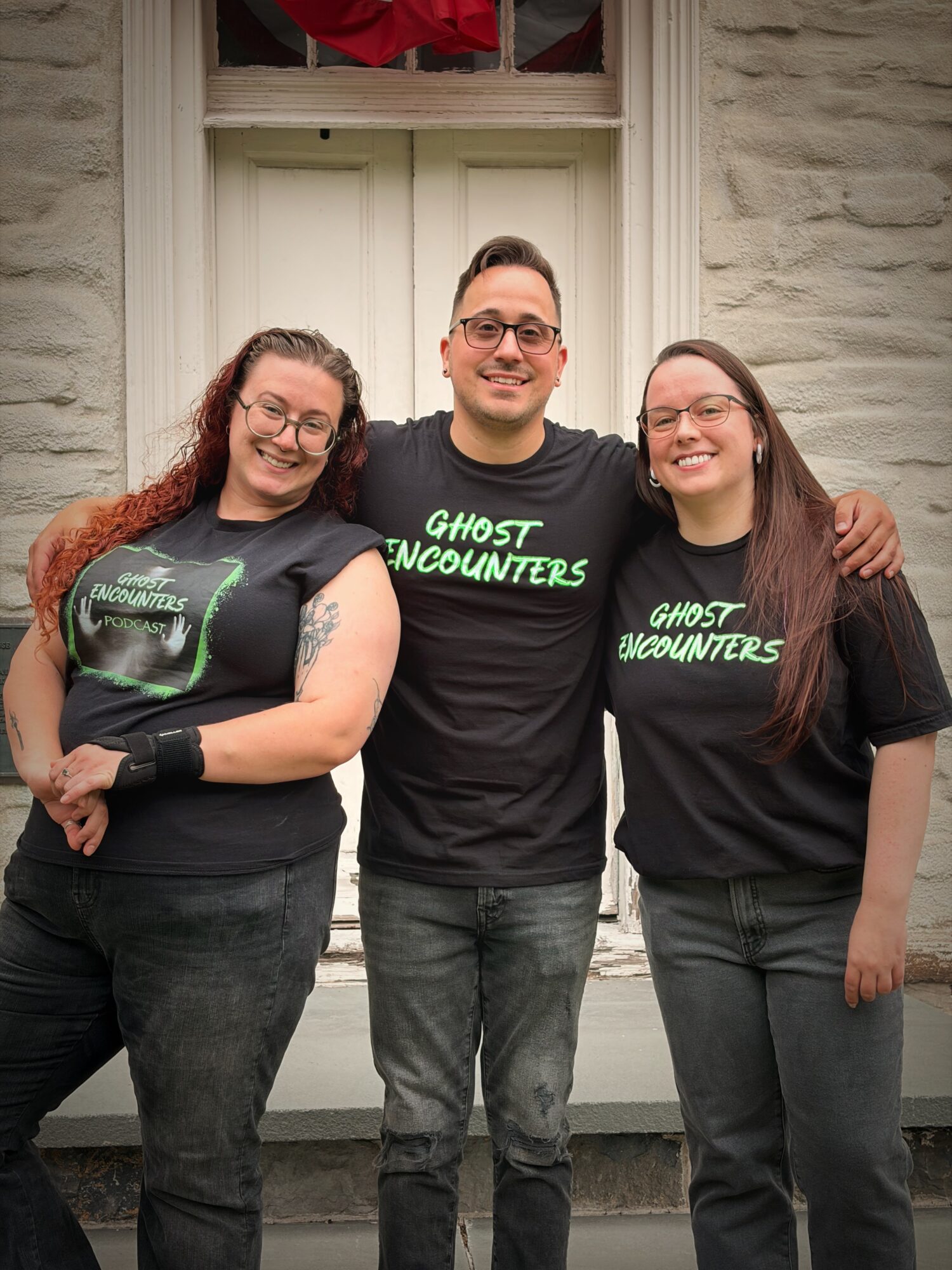 Three people standing in front of a building, smiling, with arms around each other, wearing black T-shirts with green text.