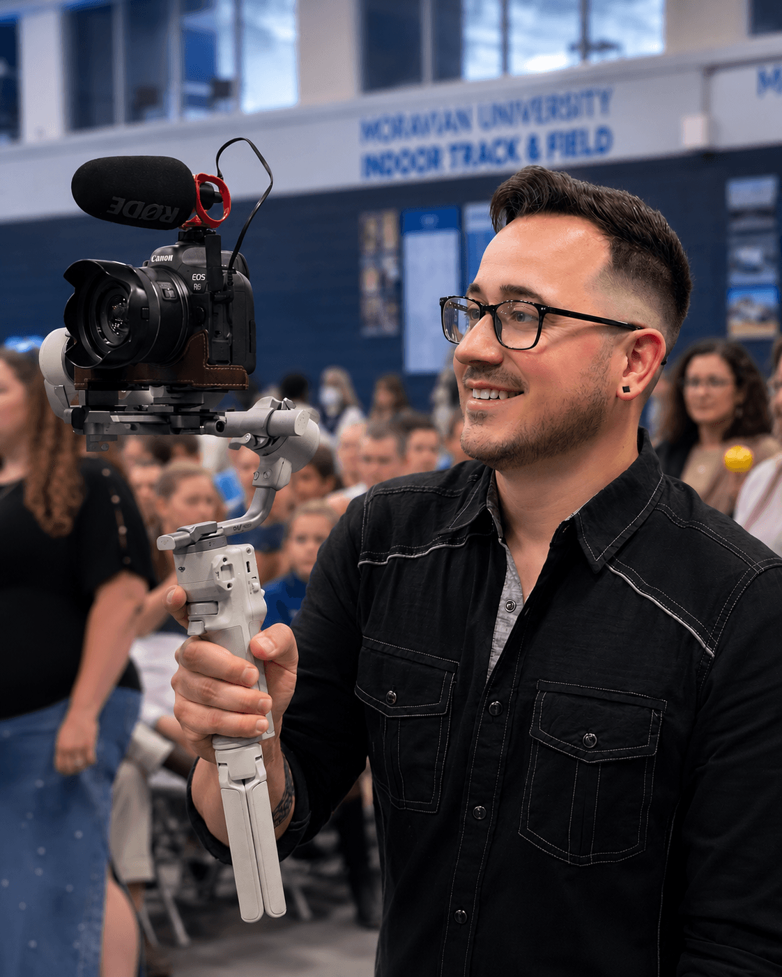 Man with glasses smiling holding a camera with a microphone in a crowded indoor space.
