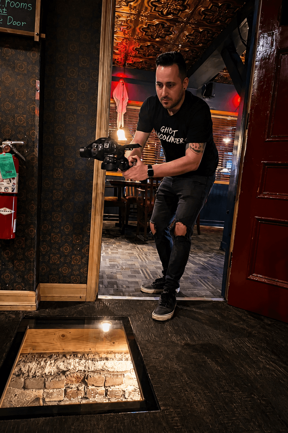 Person holding a camera, crouching in a dimly lit room with ornate ceiling, wooden door, and patterned wallpaper.