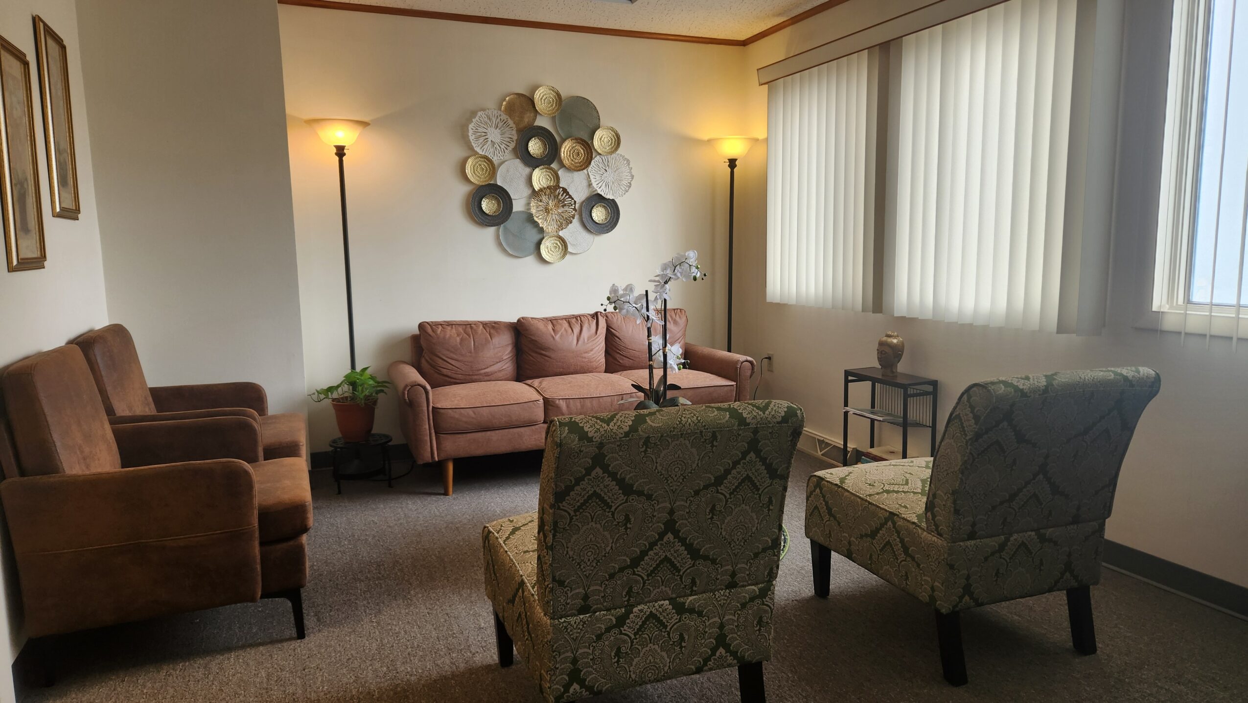 Living room with two armchairs, a sofa, and a wall decoration, illuminated by two floor lamps and natural light from windows.