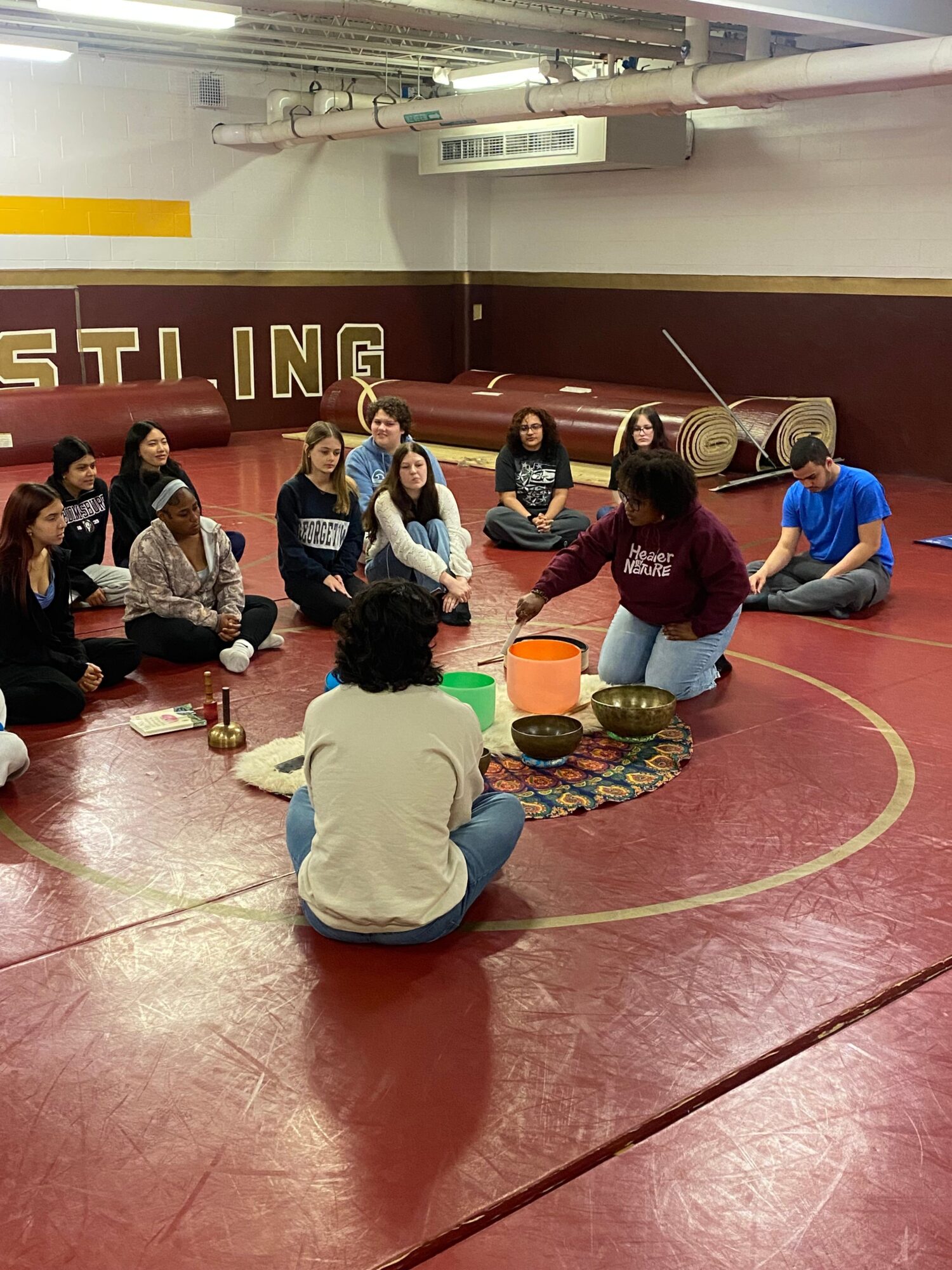 Group of children and adults sitting on a gym floor, some holding bowls, in a gymnasium with a red floor and yellow wall.
