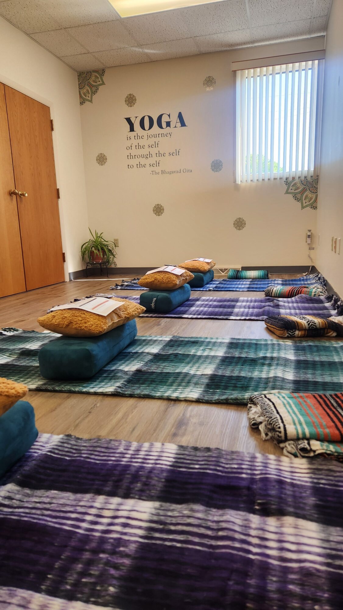 Room with yoga mats, blankets, and pillows on a wooden floor, wall with window and yoga quote, and a plant.