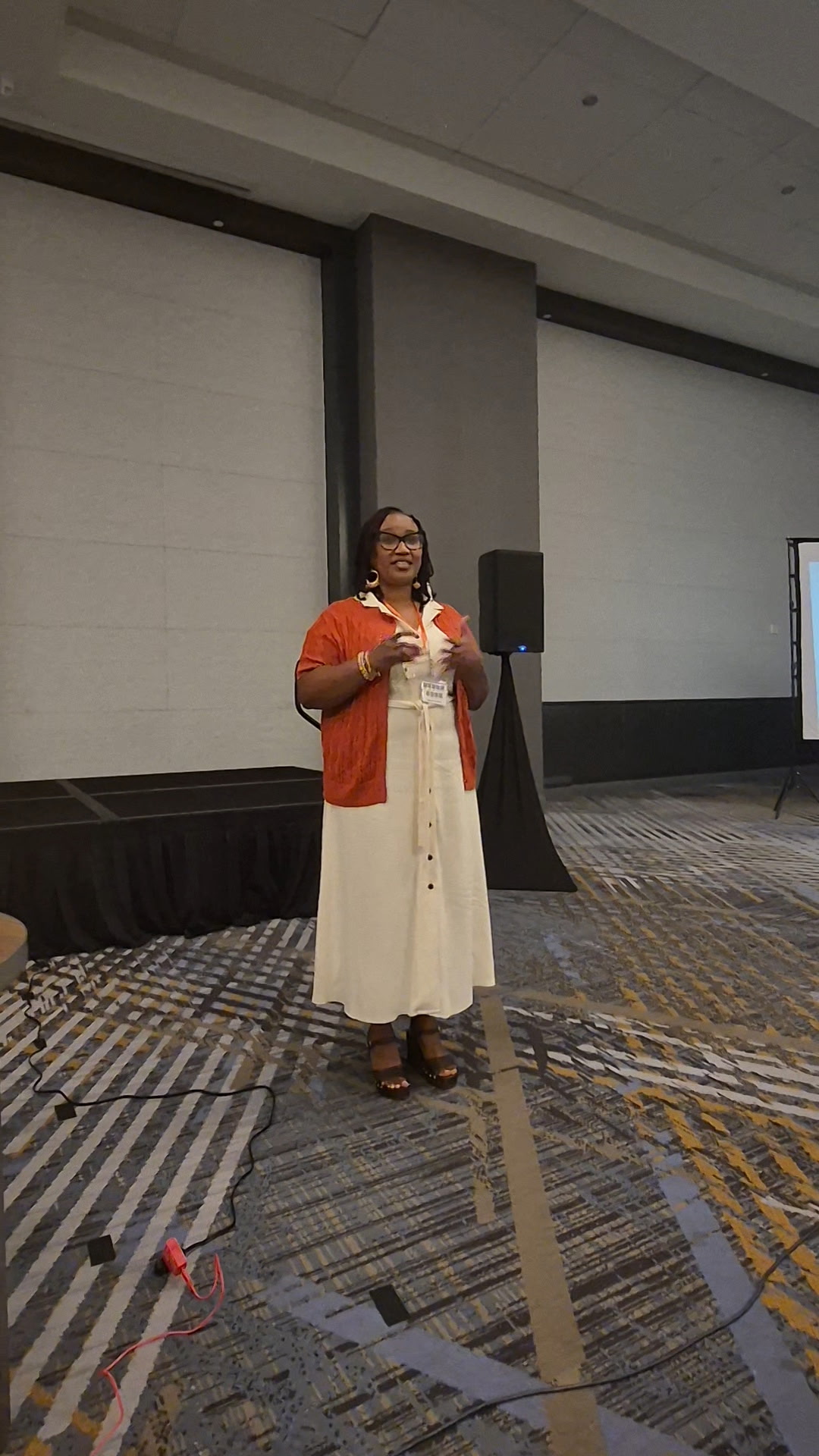 Woman standing on stage in a white dress and orange jacket, speaking into a microphone, in a conference room.