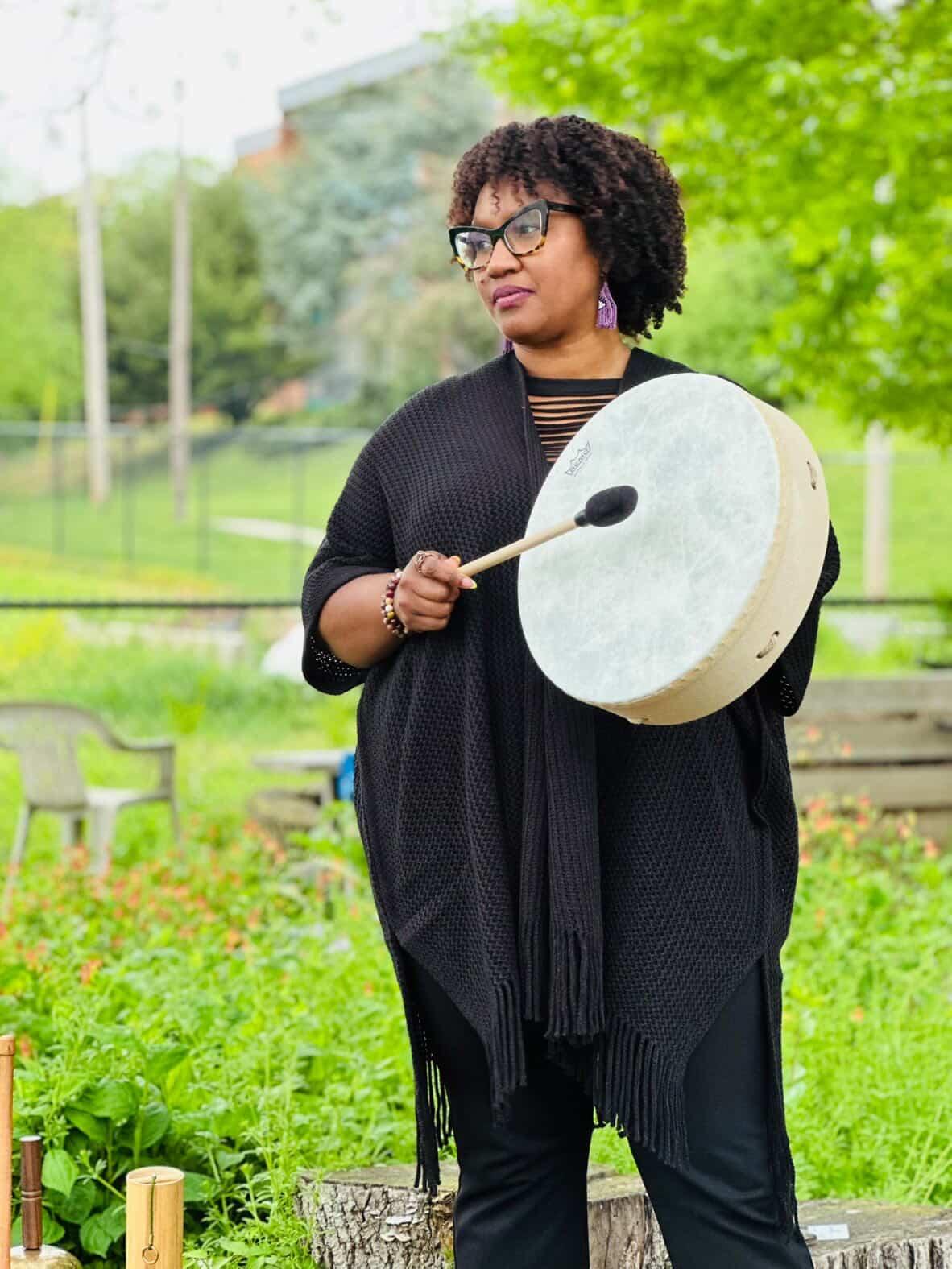 Woman with curly hair and glasses holding a large drum and drumstick outdoors in a park.