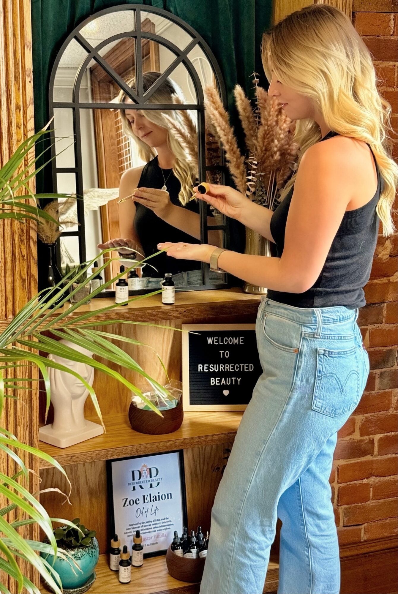 Woman with blonde hair in a black top and jeans applies makeup in front of a mirror, surrounded by plants and decor.