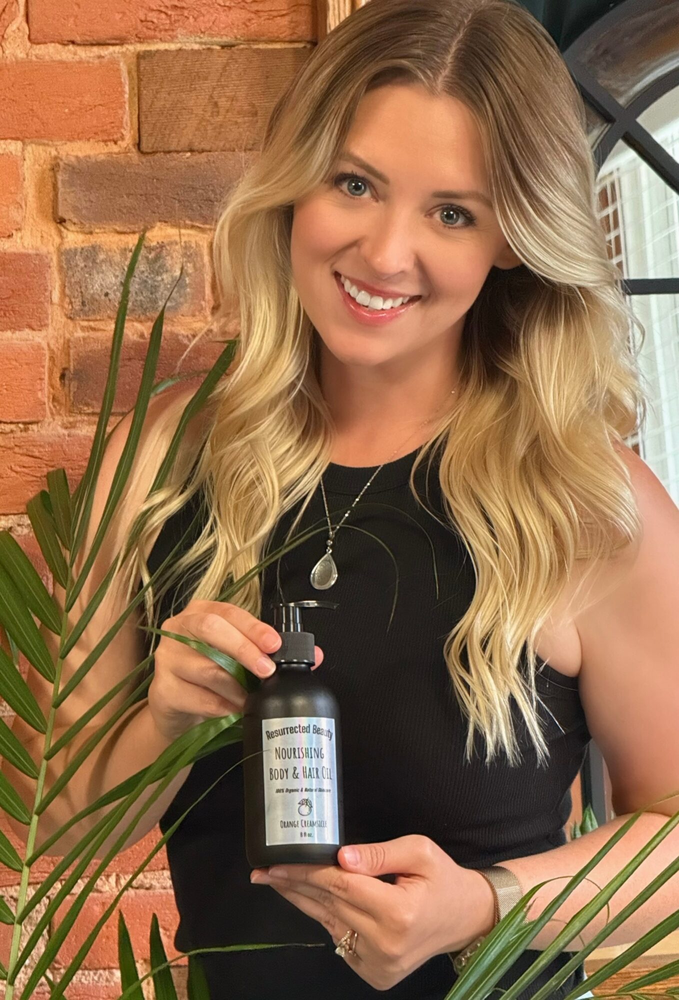 Woman with long blonde hair smiling, holding a small dark bottle, standing indoors near a brick wall and window.