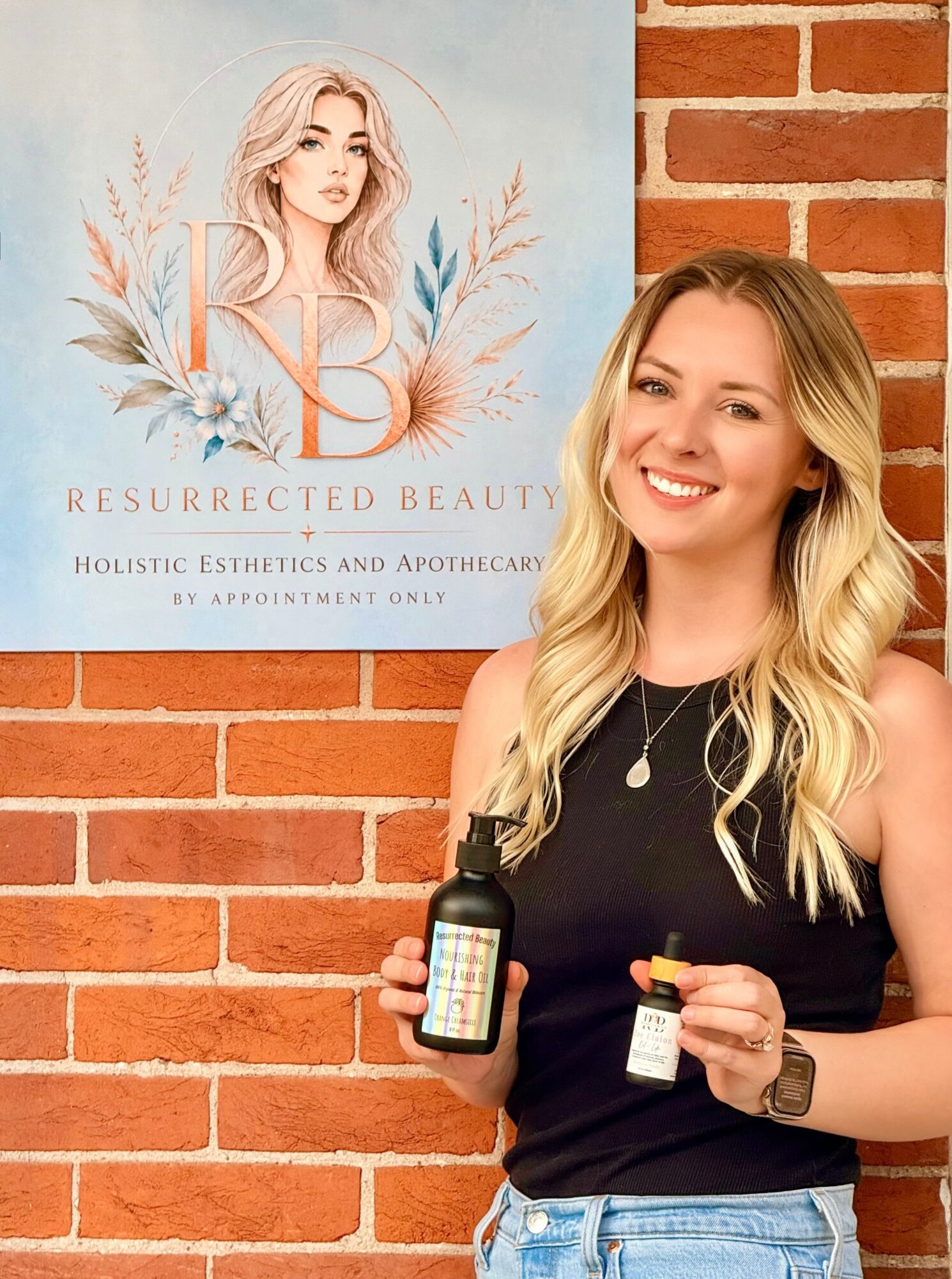 Smiling woman with long blonde hair holding skincare bottles in front of a brick wall and a poster with a woman's face.
