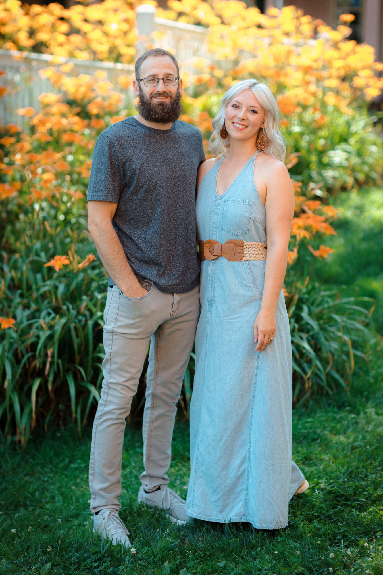 A man and woman stand outdoors in front of yellow flowers, smiling, with greenery around them.