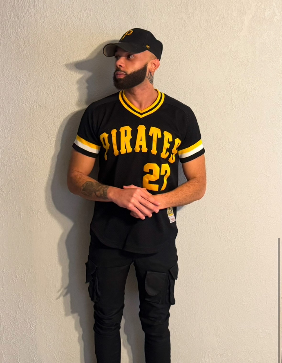 Man with beard and tattoos wearing a black baseball cap, black and yellow sports jersey, and black pants, standing against a plain wall.