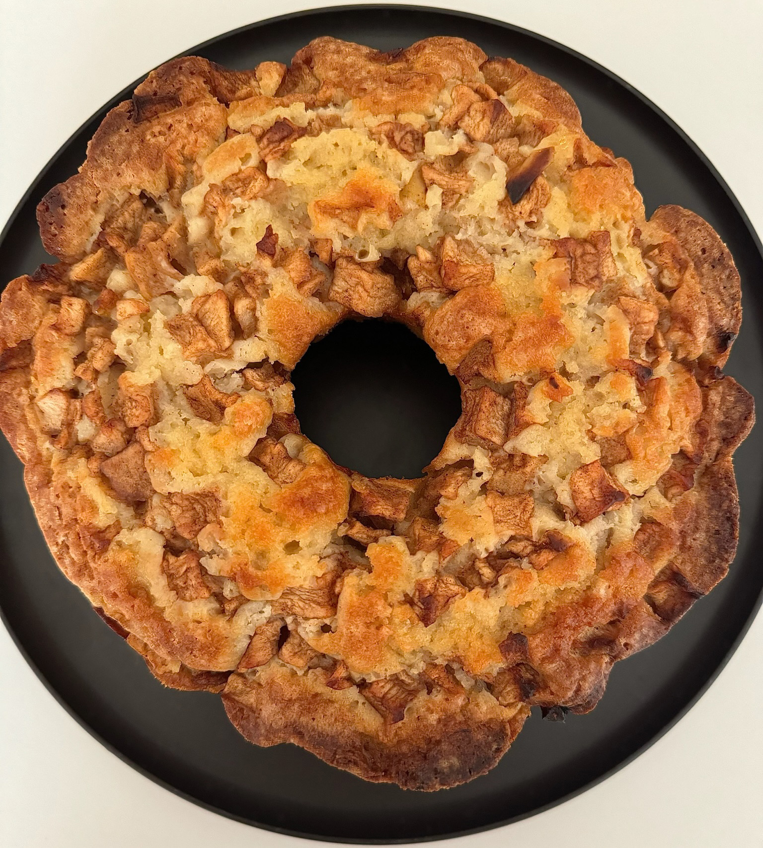 Baked apple and cinnamon cake with a ring shape on a black plate, showing a golden-brown crust and crumbly topping.