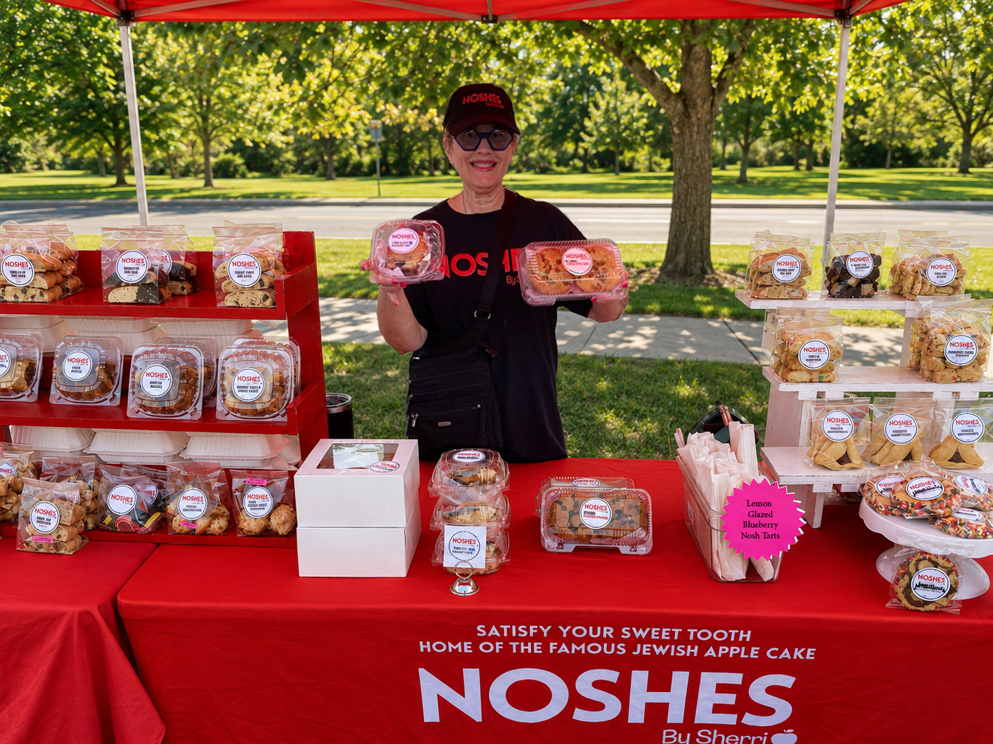 Person standing behind a table with baked goods under a red canopy in a park, holding a cake in each hand.