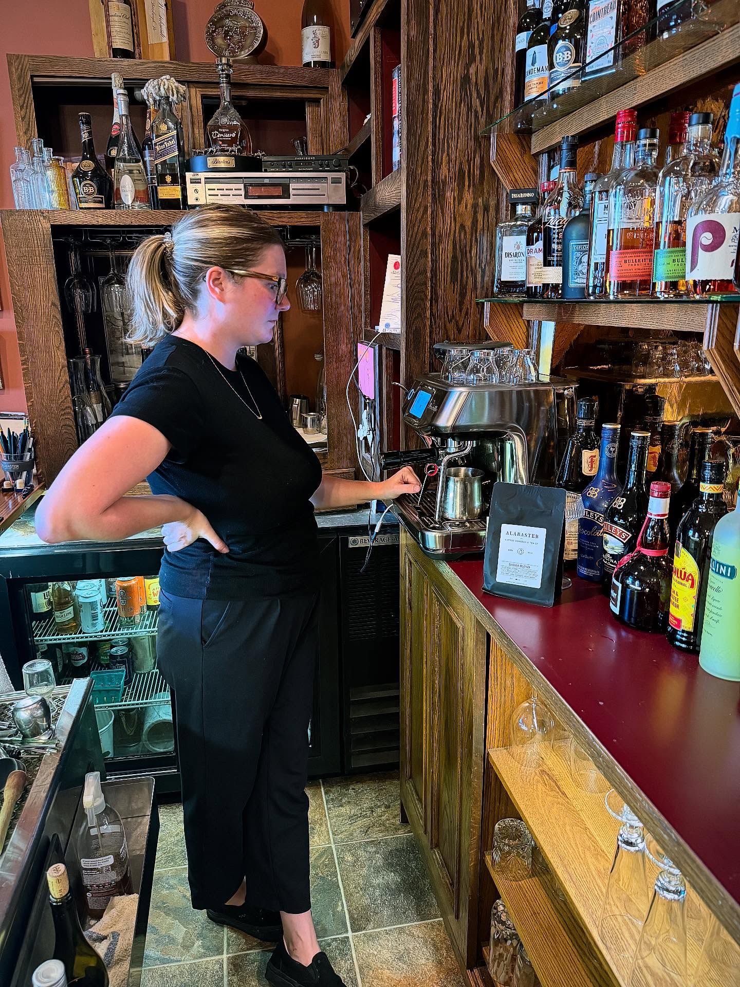 Woman with glasses and ponytail standing at bar counter, pouring drink, shelves with bottles behind her.