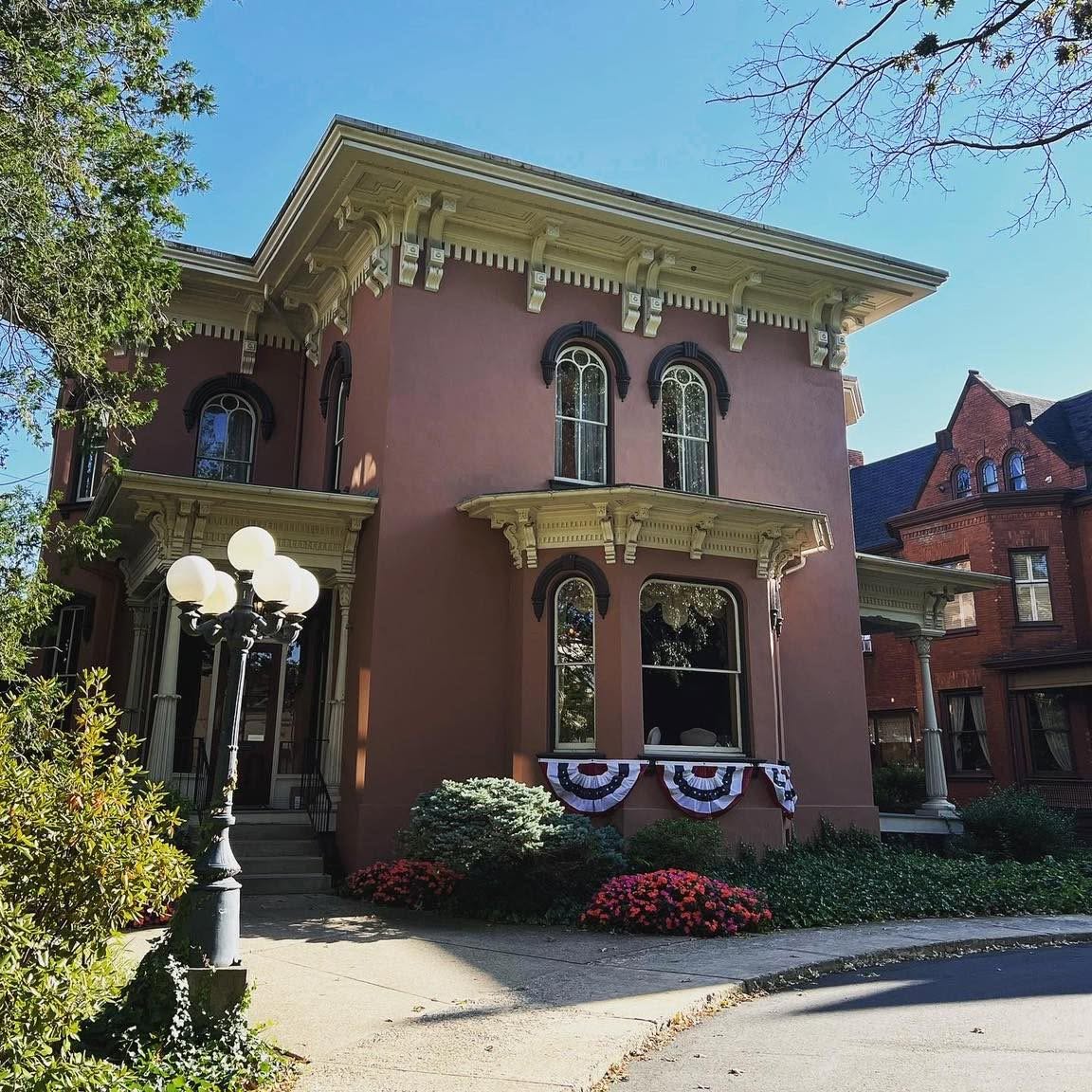 Two-story house with pink exterior, arched windows, decorative trim, and a porch with bunting, surrounded by greenery and flowers.