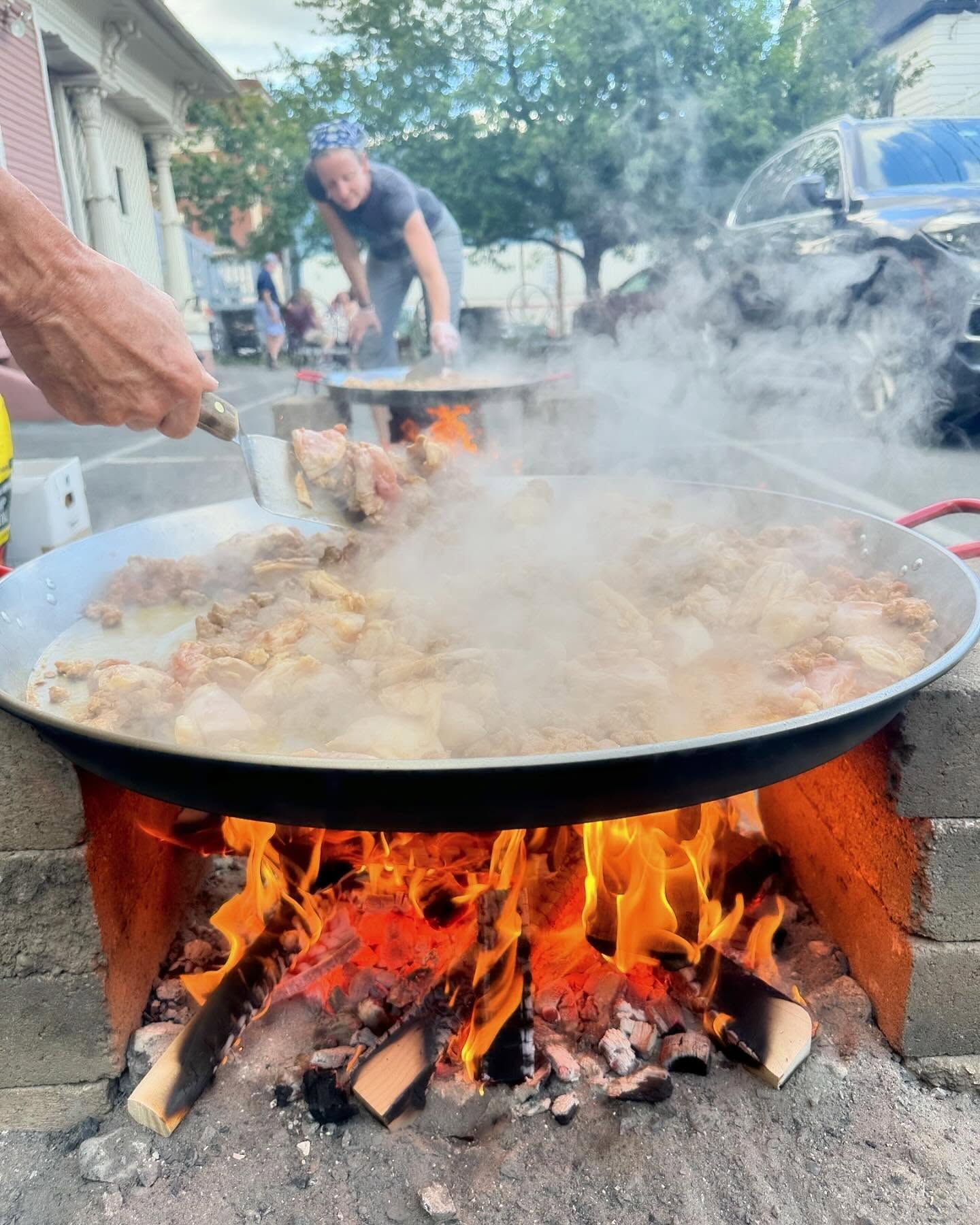 Large pan with steaming food cooking over an open flame outdoors, with a person in the background and a hand holding a utensil.