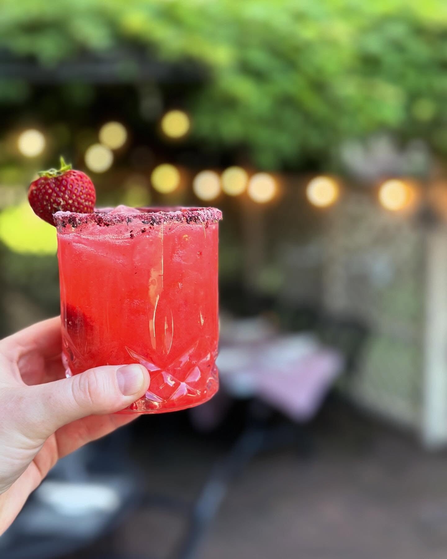 Hand holding a red strawberry cocktail with a strawberry garnish outdoors, blurred background with lights and greenery.