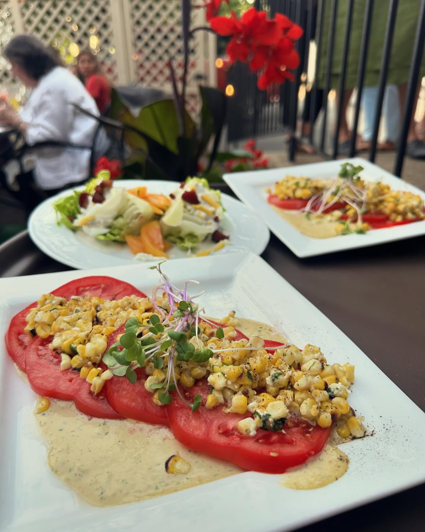 Plate with sliced tomato, corn salad, and microgreens on a table outdoors, with flowers and people in the background.