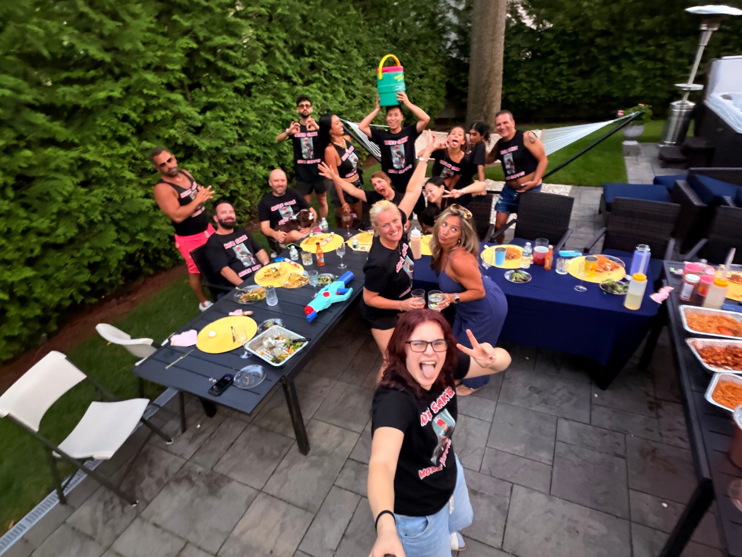 Group of people celebrating outdoors with snacks and drinks, some making peace signs, on a patio with trees in background.