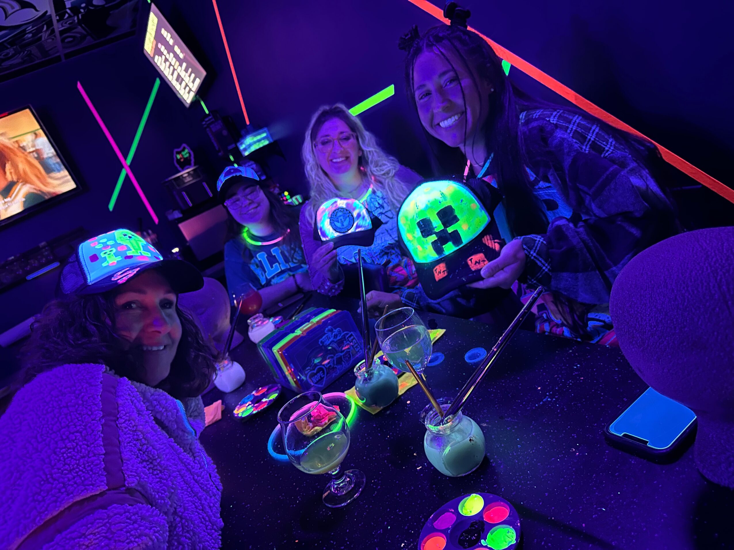 Four people in a neon-lit room holding glowing objects, with drinks and colorful decorations on the table.