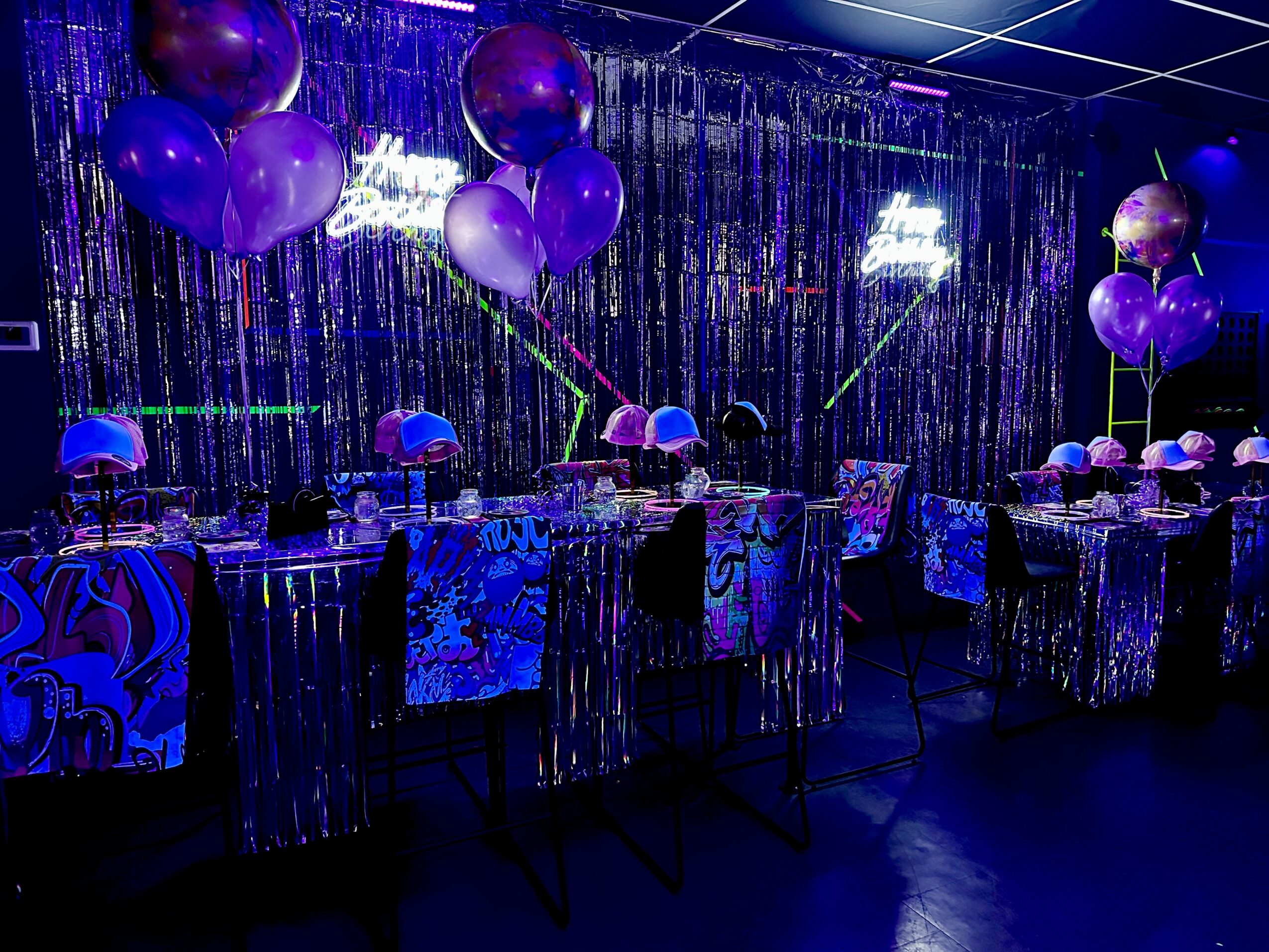 Party table decorated with purple balloons, umbrellas, and shiny tablecloths in a dark, neon-lit setting.