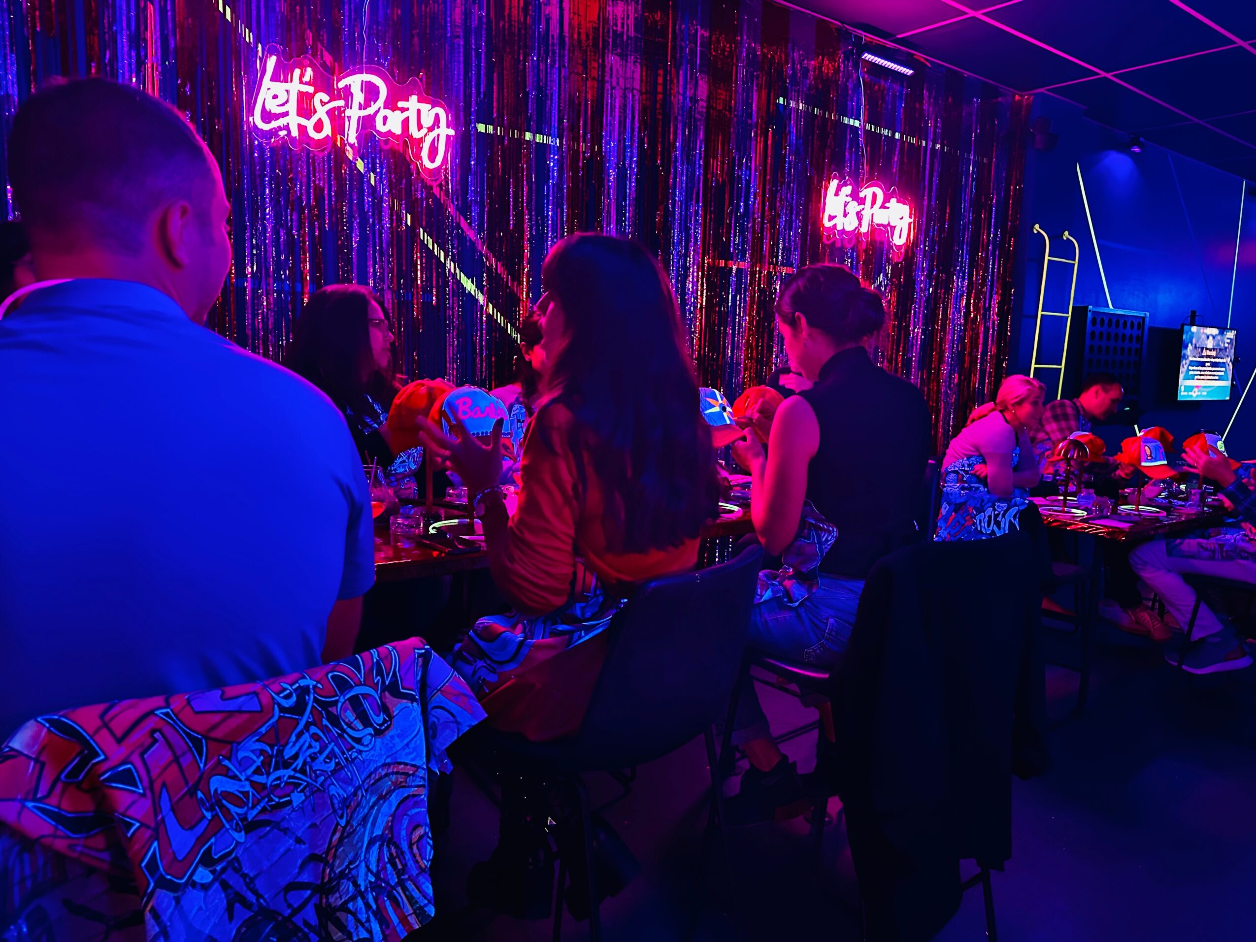 People sitting at a table in a dimly lit party with neon signs and colorful decorations.