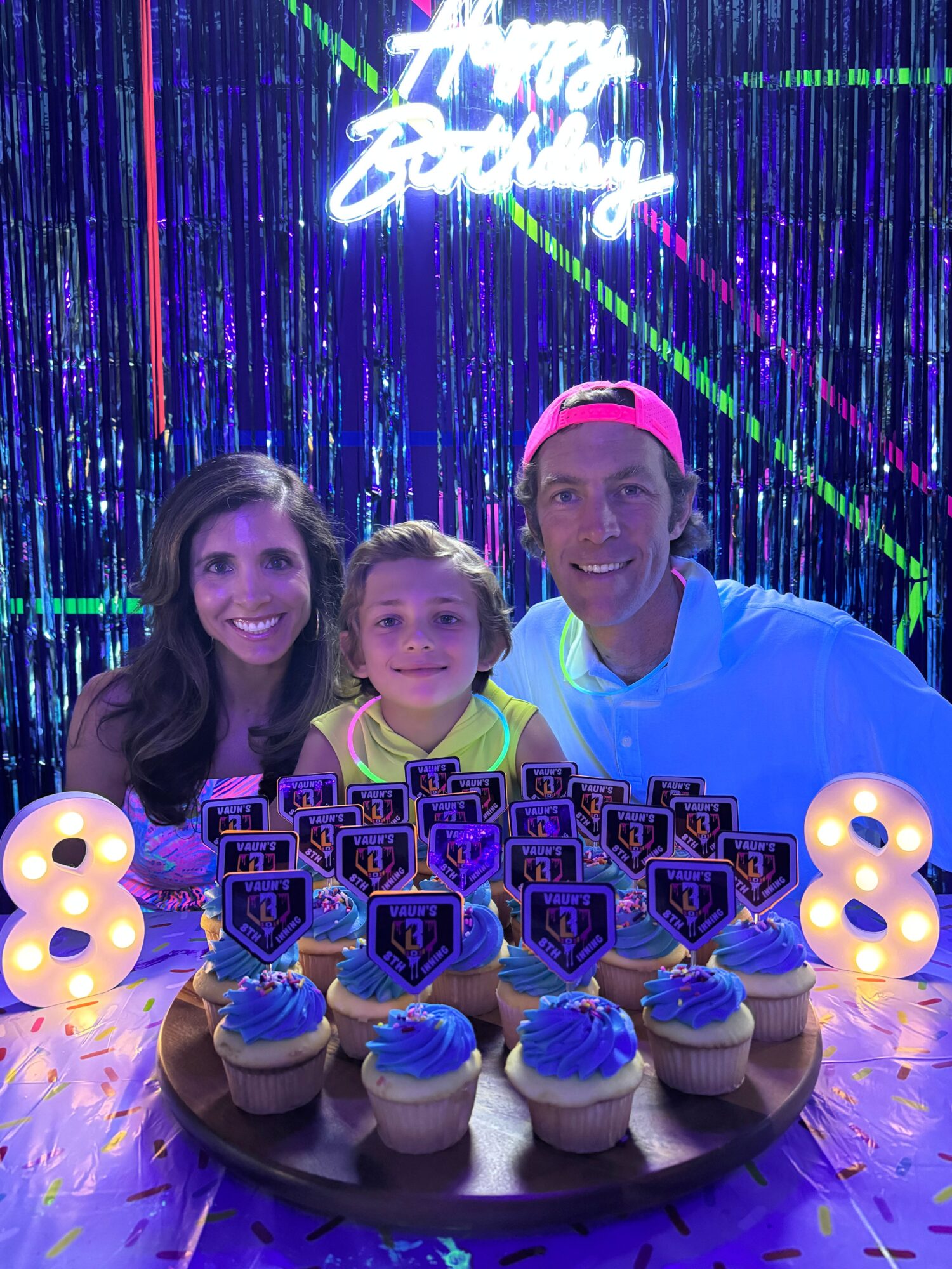Three people celebrating a birthday with a cake and candles, colorful background, and a neon sign, in a festive setting.