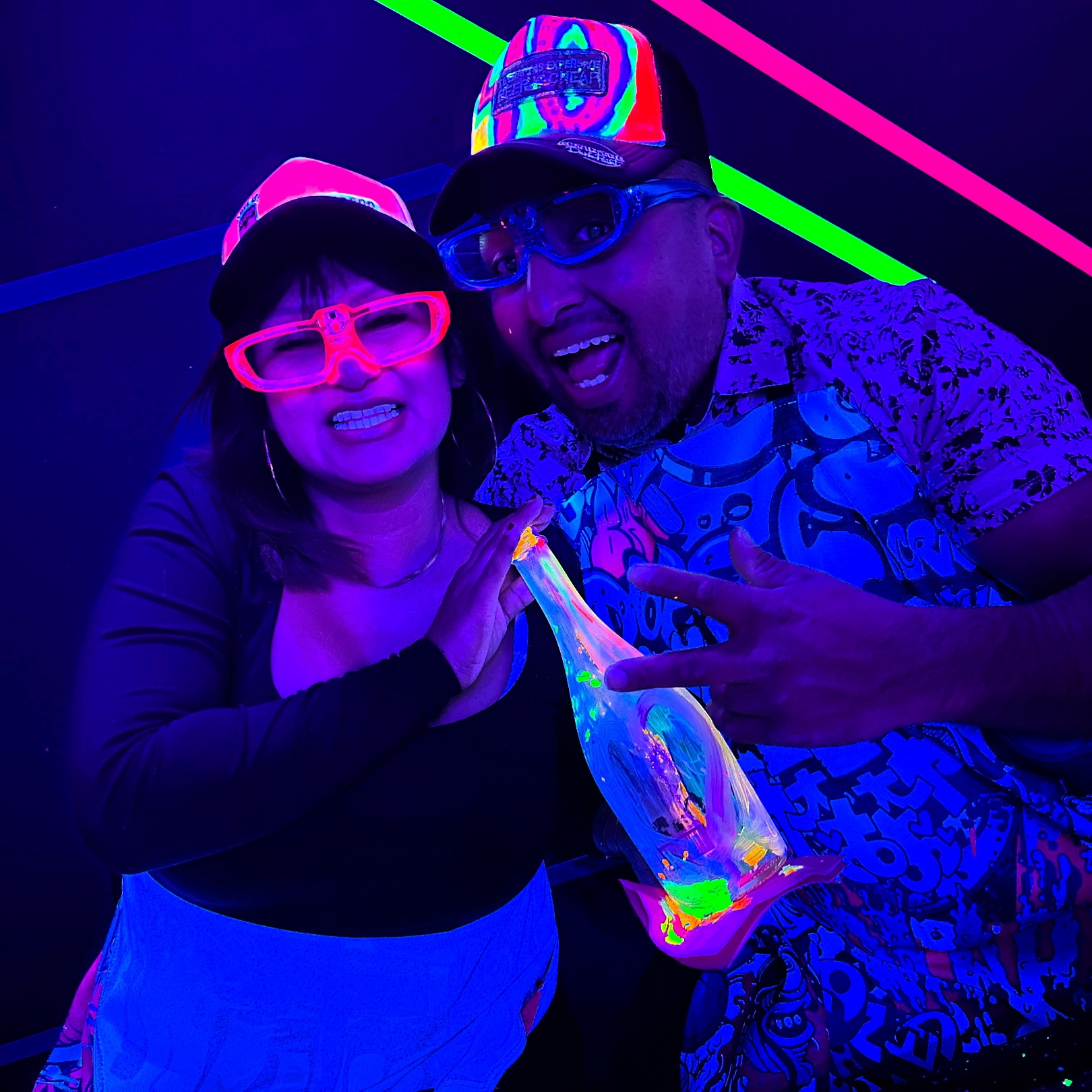 Two people smiling, wearing colorful glasses and hats, holding a glowing object in a neon-lit environment.