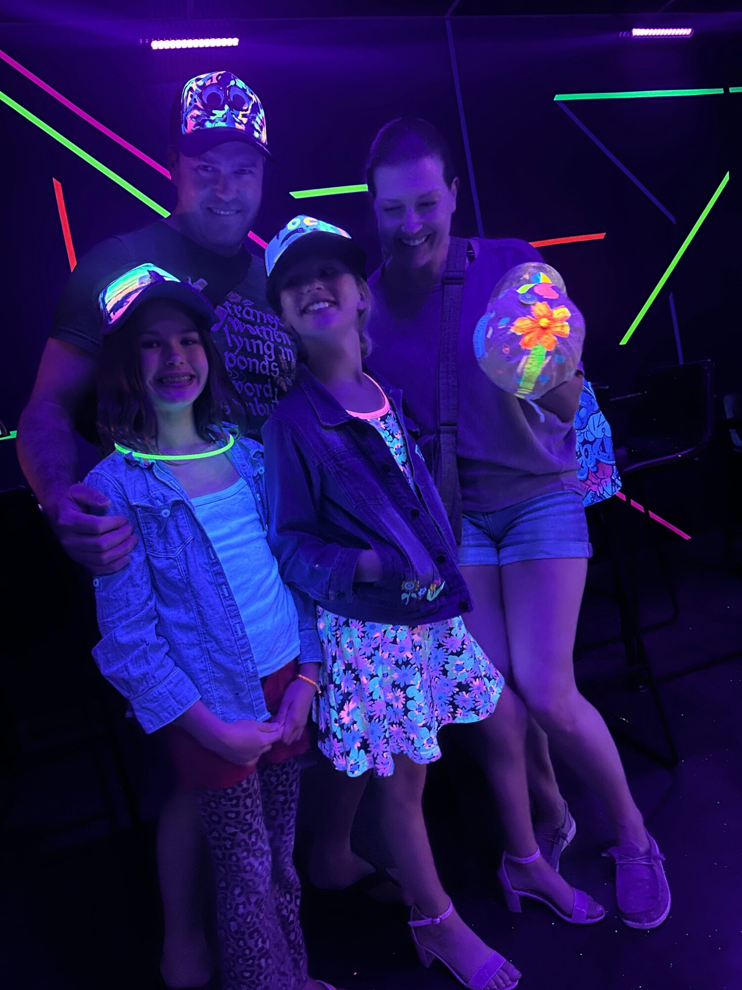 Four children in glow-in-the-dark clothing and accessories, standing in a dark room with neon lights, smiling and posing together.