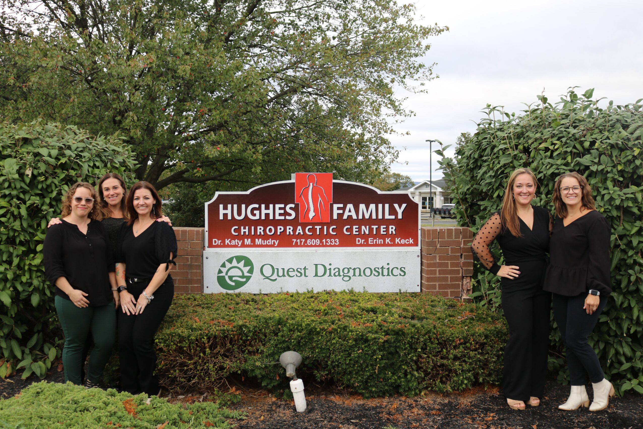 Four women standing outdoors near a sign for Hughes Family Chiropractic Center and Quest Diagnostics, with trees and bushes in the background.