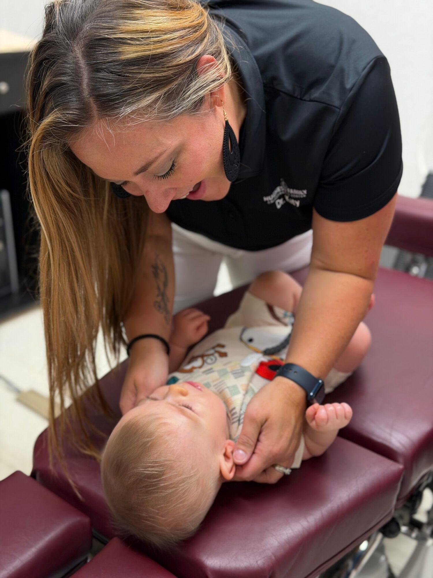 Woman leaning over a baby lying on a padded surface, holding the baby's hand and chin, in a clinical setting.