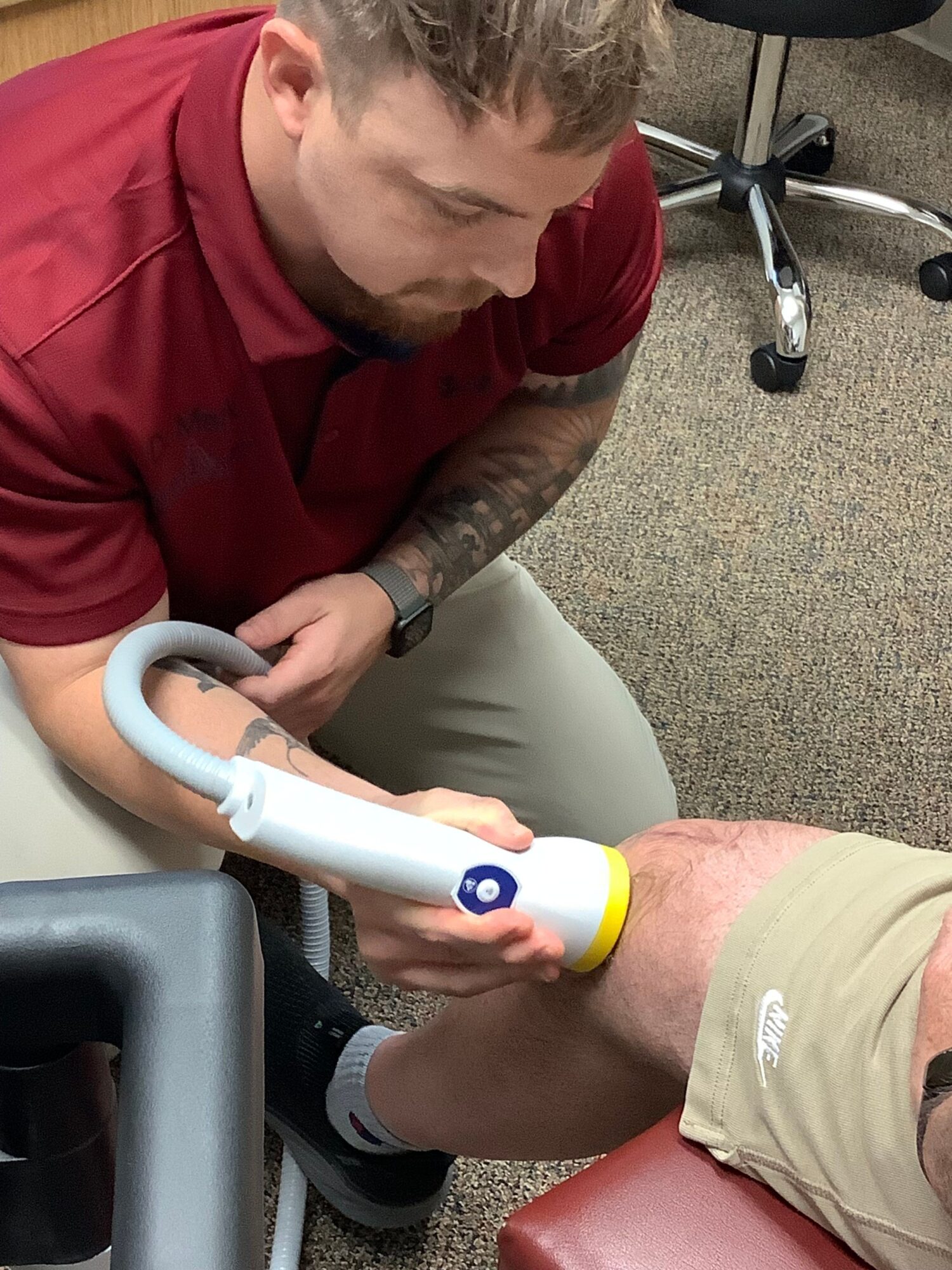 Person receiving a medical or therapy treatment with a handheld device, seated on a chair, with another person nearby.