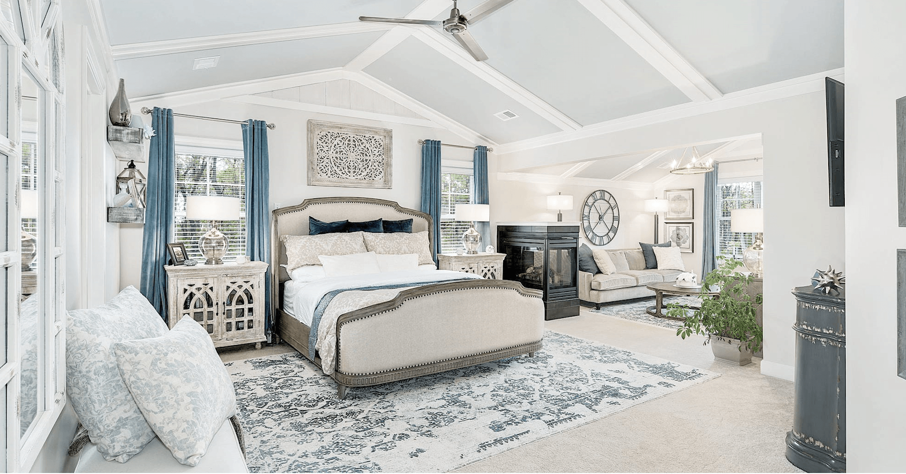 Bright bedroom with a bed, blue curtains, a rug, and seating area, decorated in light colors.