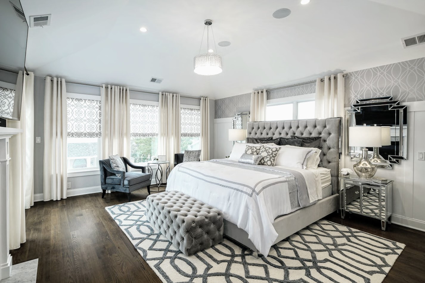 Bedroom with large bed, tufted headboard, two nightstands, armchair, ottoman, and patterned rug, with windows and curtains.