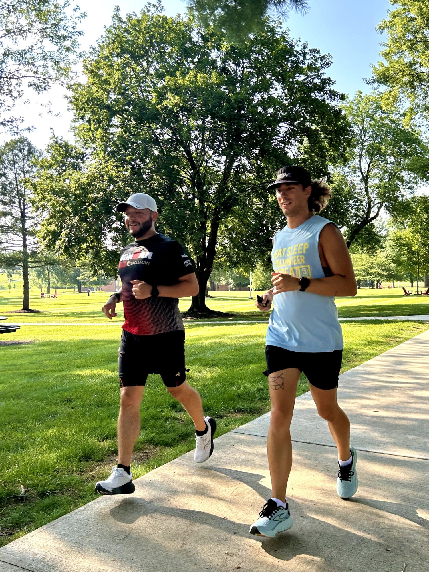 Two people jogging on a park path with trees and grass in the background, sunny weather, one wearing a cap and sunglasses.