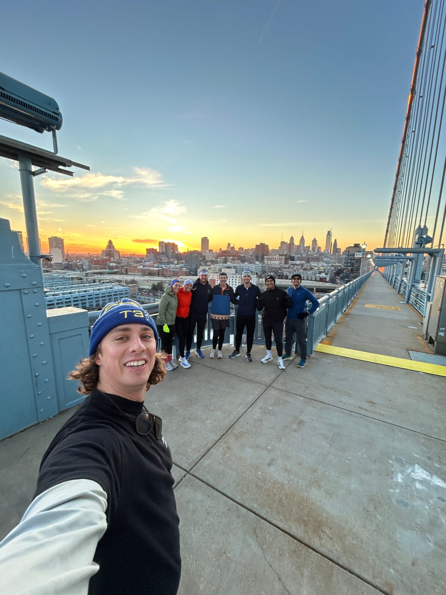 Person taking selfie on rooftop with city skyline and sunset in background, smiling, wearing black and white jacket.