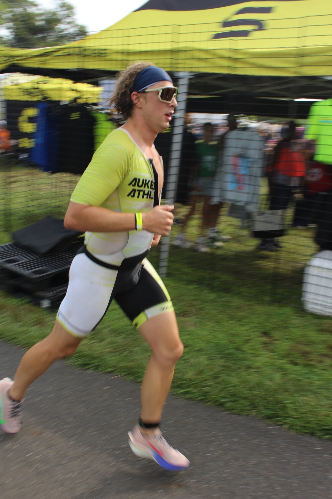 Female runner in yellow and black athletic clothing running outdoors on a paved path, with a crowd and tents in background.