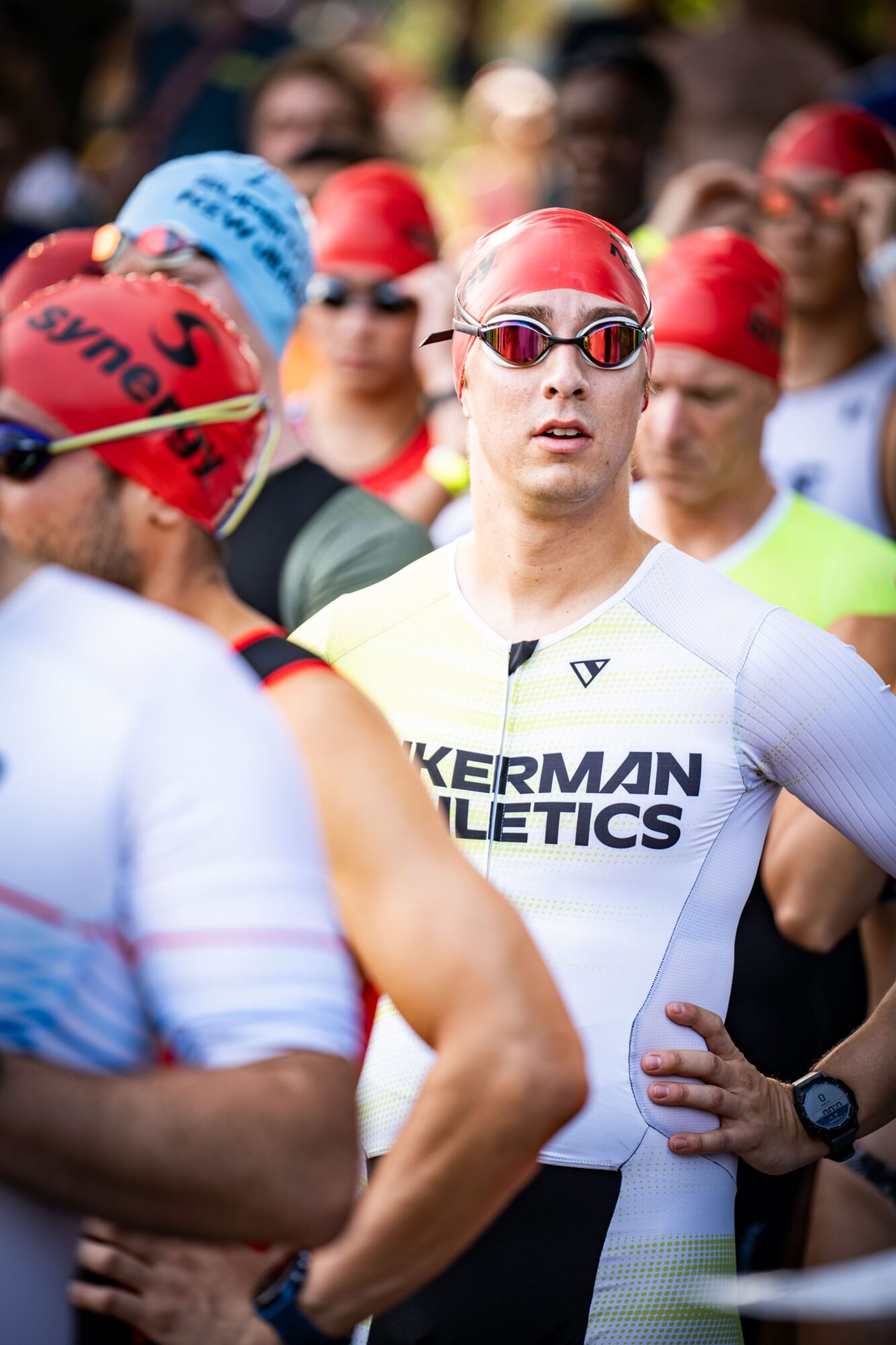 Group of athletes wearing swim caps and goggles, gathered outdoors, some looking serious or focused.